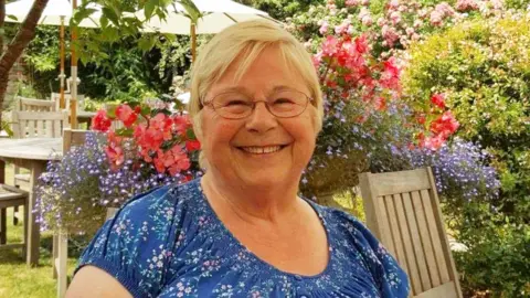 Gwynneth Exall wearing a blue and floral top, wearing glasses and with short grey hair, smiling at the camera, while sitting in a garden