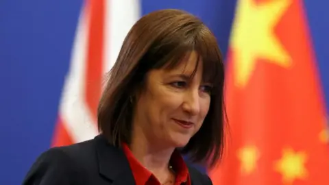 Reuters UK Chancellor Rachel Reeves dressed in a black jacket and red shirt standing in front of a backdrop of two flags representing China and the UK