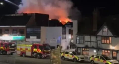 Fire vehicles parked on a road with a fire in a building behind.