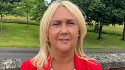A woman with shoulder length blonde hair looks into the camera. She is wearing diamond earrings and a red jacket with a white top. She is standing in front of a low wall and in the background is a green area with trees.