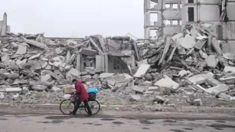 Getty Images Un homme marche avec un vélo portant des sacs remplis d'effets personnels à côté d'une rangée de bâtiments réduits en tas de décombres à Myrnohrad, dans l'oblast de Donetsk, dans l'est de l'Ukraine.