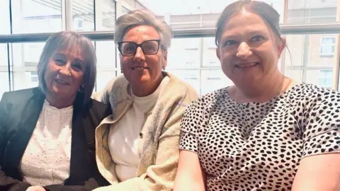 MICK LUNNEY/BBC Three women sit indoors and pose for a photograph. They are Tracy Watson, Joanne Heald and Nicola Lovell.