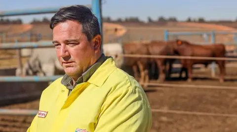 Gallo via Getty Images John Steenhuisen, dressed in a yellow overall jacket, is seen standing in front of cattle at a farm