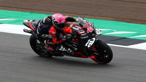 Asprilia motorbike covered in sponsor names. It has some red trim. The rider has a pink helmet and red and black leathers. The bike is leaning to the right as the bike takes a right-hand bend on grey tarmac, with a green and white strip to the biker's left.