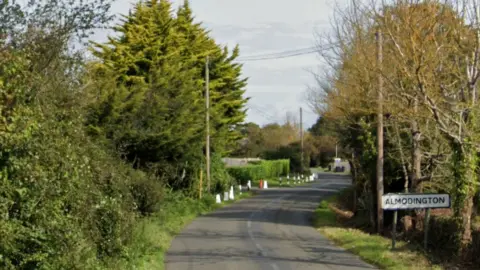 Google Maps Almodington Lane in West Sussex near Chichester. Pictured is a narrow lane with green trees on either side and a white sign which reads Almodington