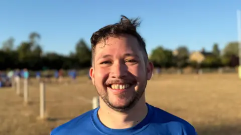 Elliot Deady/BBC A man - Jonathan Howes - smiles straight at the camera while standing on a rugby pitch. Several players are behind him in the distance.