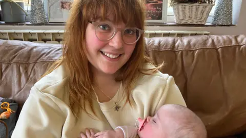 A woman with auburn hair with a fringe, who is wearing round, beige glasses and a yellow, round-necked jumper. She is holding a sleeping baby in her arms. She is sitting on a brown, leather sofa.
