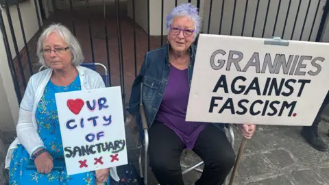 Two women sat in chairs holding placards. One reads "Grannies against facism" whilst the other reads "heart ur city of sanctuary".