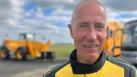 A man looks towards the camera wearing a yellow and black driving suit with the "JCB" logo on the front.