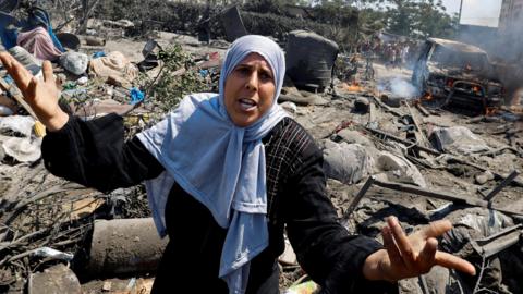 A woman raises her arms in anguish. Behind her is the aftermath of an Israeli airstrike, including a burnt out truck.