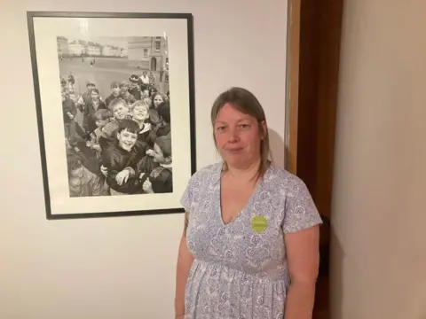 Becky Smith is in a grey patterned dress. She has long blonde hair and is standing in front of a black and white photo.