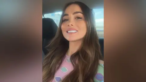 Jade Morgan Jade is sat in a car and is smiling at the camera. She is wearing a pink top with green and purple spots.