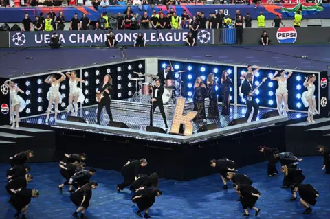 Live Nation/ Getty Images Still image of The Killers performing in Budapest with dancers off-stage in black outfits to match the band and contrasting white outfits of on-stage dancers.