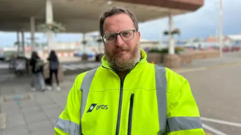 BBC A man wearing a high vis jacket with DFDS logo on the right chest area, background of the harbour is blurred.