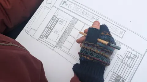 Ric's hand in a glove drawing a shop front in pencil.