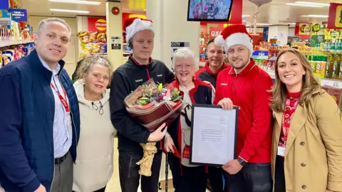 Three women and four men huddle in a supermarket. One man is wearing a red shop uniform, santa hat and holding a framed letter. Another man is in a black uniform jacket, santa hat, headset and holding a bunch of flowers. Shelves of food can be seen behind the group.