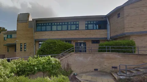 Photo of Shrewsbury Crown Court; it's a modern building, built out of sand-coloured bricks and grey-black-framed windows. In the foreground you can see bushes and shrubs, and steps and hand railings leading up to the court entrance. It's a sunny but overcast day. 