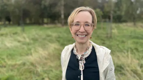 Louise McManus has short blonde hair. She wears round glasses, a navy sweater with a blouse tied with a bow at the neck, and a cream waterproof jacket. He smiles widely at the camera and stands in the field. There are a few small trees behind him.