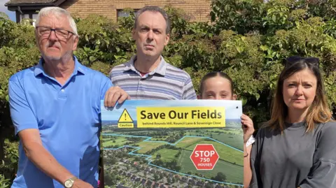 Malcolm Lines is wearing a blue t-shirt and has short white hair and glasses, he is standing next to rob jones in a stripey t-shirt, next to him is grace who has her hair tied uo and they are all holdinhg up a save our fields poster. Next to Grace is her mum Stacy who has shoulder-length light brown hair and is wearing a grey t-shirt