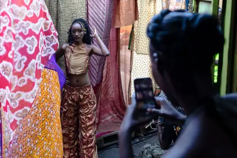 TONY KARUMBA / AFP / GETTY IMAGES A woman wearing a bronze-coloured top and trousers stands next to draped pieces of colourful fabric while having her photo taken.