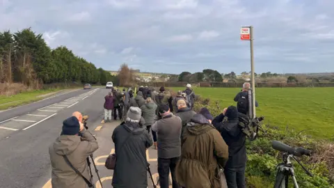 A group of bird watchers in a bus stop, all looking through telescopes or binoculars in the same direction, with a road beside them