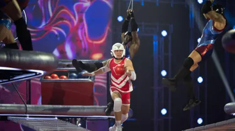 BBC/Hungry Bear Media Ltd/Graeme Hunter Mark Billings is running across a wobbly walkway, suspended in the air, as Gladiators swing in from the side while dangling from rings, to try to knock him off.