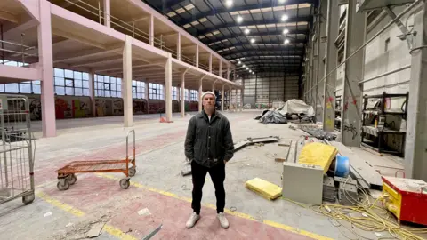 BBC A man in a black jacket, black trousers, white trainers and a white woolly hat stands in a huge foundry. There are hardware trollies, tools, building materials and equipment covered by tarpaulin. To the left, a newly built mezzanine structure. The huge building is well lit and covered by a new roof. There is graffiti on the far wall beneath the mezzanine.