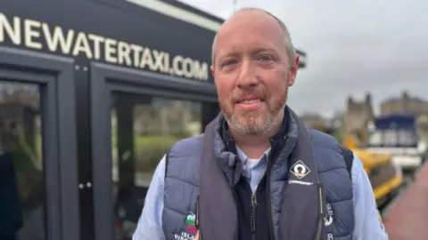 BBC A man stands on a jetty beside a water taxi. He has a beard and short grey hair. He is dressed in a blue shirt, navy very and blue waterproof gilet and safety vest. The background is blurred, but there are boats in the background and he is standing beside an office with a sign that reads 'NEWATERTAXI.COM'