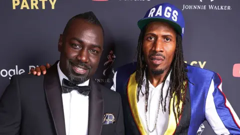 Getty Images DJ Fonti and MC Bushkin at a party, with Fonti wearing a black tuxedo, white shirt and black bow, and Bushkin wearing a blue coloured blazer and a hat with "RAGS" written.