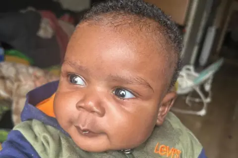 A photo of baby Akram, with big grown eyes and short brown hair, wearing a green and blue Levi's jumper. He is looking to the left of the camera. 