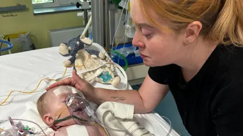 Kristy Evans A baby and a mother looking over him. The mother has a black t-shirt on and ginger hair. The baby lies in the hospital bed and has tubes on his face and body.