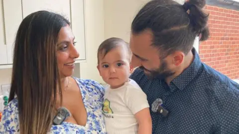 A mum, dad and baby are pictured. The baby is in the middle, smiling. The mum is on the left wearing a blue floral top. Dad is wearing a dark blue shirt with white spots. Everyone is smiling.