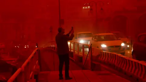 A man takes a picture while cars drive with their headlights on in red air.