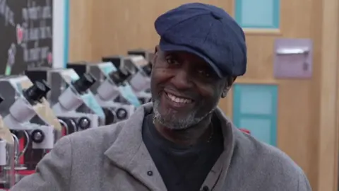 A man in a flat blue cap and grey jacket with a black jumper underneath is smiling at the camera. On his left behind him there is a row of microscopes