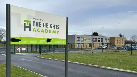 There is a white and green sign at the school entrance at the Heights Academy in Smith's Wood. The background has a two -storey building that provides the Hasa flat roof and a service road. A car park can also be seen in front of the building. 