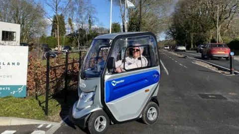 Dorset Council A smiling man in a covered blue and grey mobility scooter alongside a newly opened cycle path separated from the road alongside.