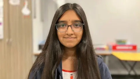 Eyva is smiling and looking at the camera. She is wearing a navy hoody and red and white T-shirt. In the background there are tables and cupboards inside a classroom. 