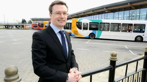 Stagecoach Stagecoach East Midlands Managing Director Matt Cranwell standing to the left of the photo. He is middle-aged wearing a black blazer, light blue shirt and navy tie with white spots. He is smiling, has dark hair and is wearing silver thin framed glasses. He is in a coach park, with a row of yellow, white and turquoise single decker buses behind him.
