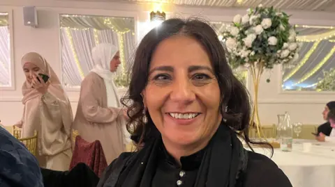 Nicola Haseler/BBC A woman smiles happily at the camera while sitting at one of the tables. She wears a black blouse and scarf, and her head is uncovered. Behind her other women mill about. 