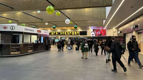 People inside Leeds railway station walking to the ticket barriers with the information desk and departure boards in the background