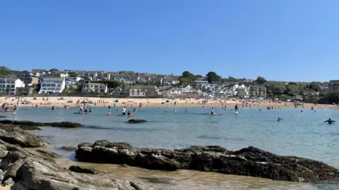 People enjoying the beach at Porth near Newquay in Cornwall on a sunny day. Dozens of people are out on the water, including several on paddleboards.