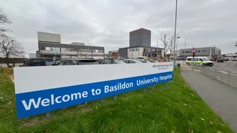 A general view of the entrance into Basildon Hospital. A blue and white signs reads the hospital's name. A car park can be seen behind it along with hospital buildings. 