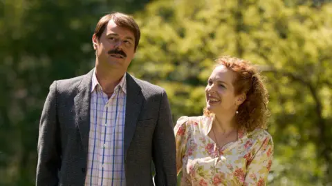 Robert Viglasky/Disney+ The characters Freddie Jones and Lizzie Vereker walking arm-in-arm. Freddy has brown hair and moustache, wearing a dark grey blazer and blue check shirt. Lizzie has curly red hair, wearing pearl drop earrings and necklance and a floral dress.