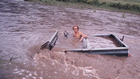 Oriya Douglas-Hamilton. An old photograph of Ian Douglas-Hamilton sitting in an open-top Jeep-style vehicle that was almost completely submerged in a brown river. 