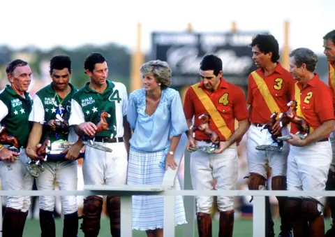 Princess Diana Archive / Getty Images The prince and princess stand next to each other as other polo players dressed in polo gear stand alongside them, holding equine trophies with a billboard in the background advertising Cadillac.