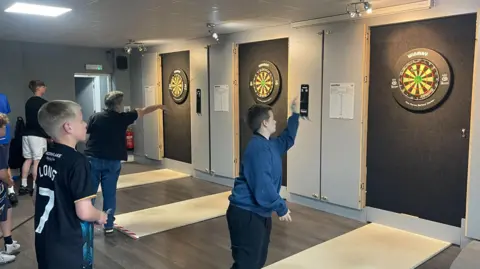 Three darts boards on a wall with young players waiting to throw darts at them