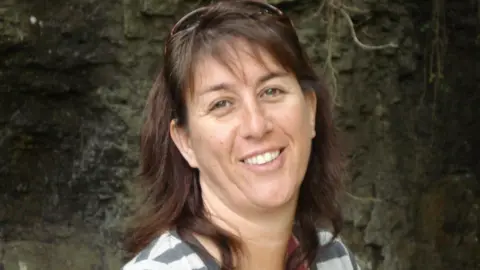 Stephen Price Donna, who has shoulder length dark-brown hair smiles at the camera. She is outside, and appears to be standing in front of a rocky cliff or cave. She wears a striped white and grey top. 
