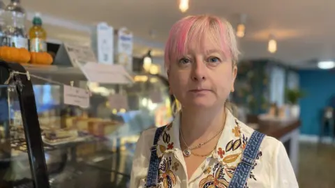 Michelle Langan, who has blonde hair cut into a fringe with pink highlights, poses for a picture in a café wearing a flowery white shirt with denim overalls.