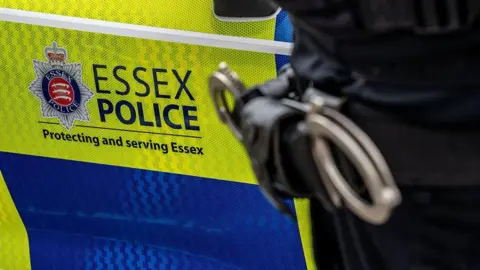 Close-up view of Essex Police vehicle's door. The force's logo, on a neon yellow and blue chequered background, can be seen with the words "Essex Police Protecting and serving Essex". A pair of handcuffs on an officers hip can be seen to the right of the door.