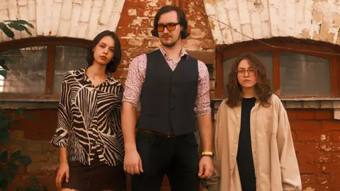 Polina Grebenik  Two women stand on either side of a tall man in front of a redbrick building. The man wears a button-up vest over a floral patterned shirt, sleeves rolled up. His combover hairstyle and sunset-tinted glasses complete his 1970s look. The woman on his left has a short black bob haircut and wears a blouse with black and white zebra print. The woman on the man's right has shoulder-length, curly hair and wears glasses. She's wearing a cream-coloured, oversized shirt over a black outfit.